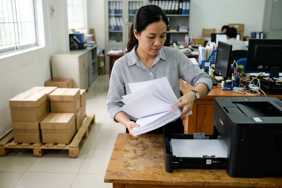 Nhân viên quạt tơi xấp giấy trước khi nạp máy in, thùng giấy kê trên pallet gỗ phía sau
