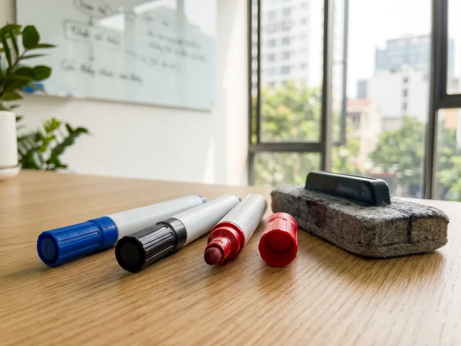 Bốn cây bút lông bảng và bông lau bảng đặt trên bàn trước bảng trắng, một cây mở nắp lộ đầu ngòi